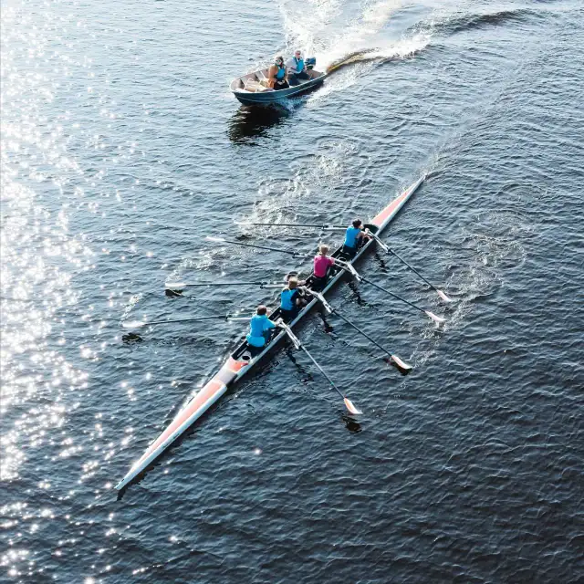 A multi-person rowing team in a sleek racing boat on open water, with a coach boat following behind; sunlight glints on the water and oars are in sync as the crew rows.