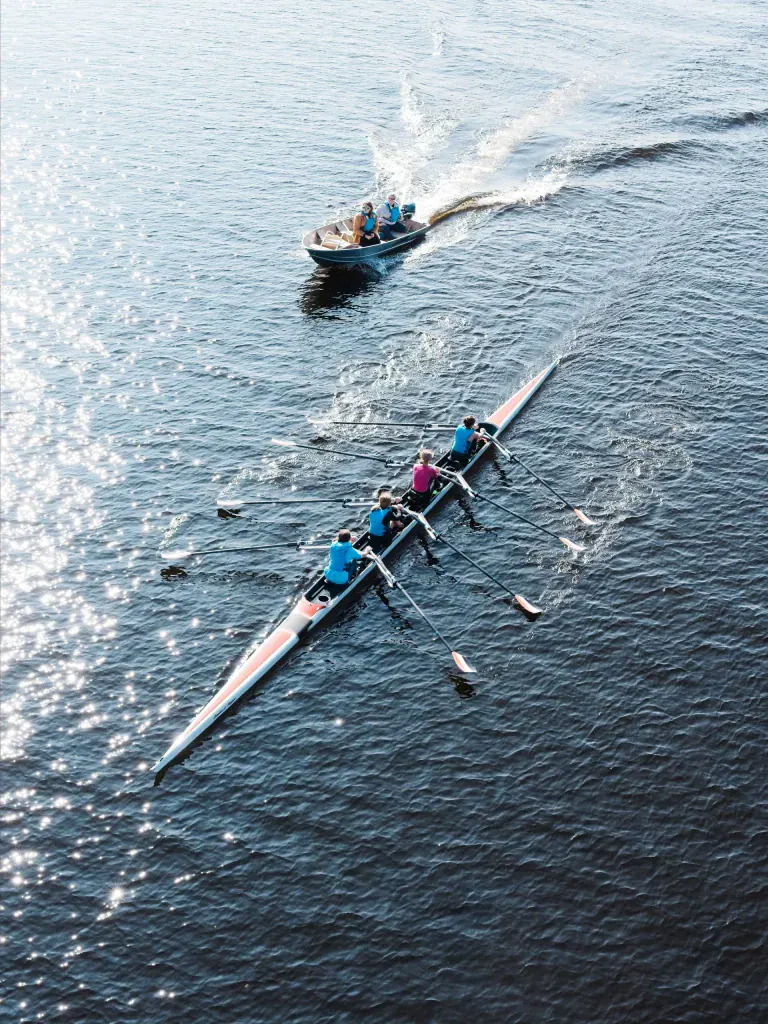 A multi-person rowing team in a sleek racing boat on open water, with a coach boat following behind; sunlight glints on the water and oars are in sync as the crew rows.