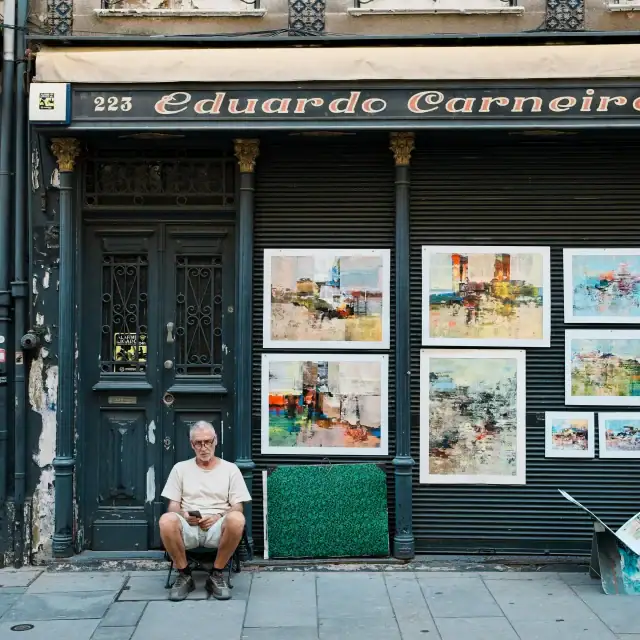 Older man sitting outside an art gallery with framed paintings displayed in the storefront window.