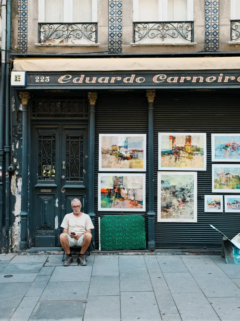 Older man sitting outside an art gallery with framed paintings displayed in the storefront window.