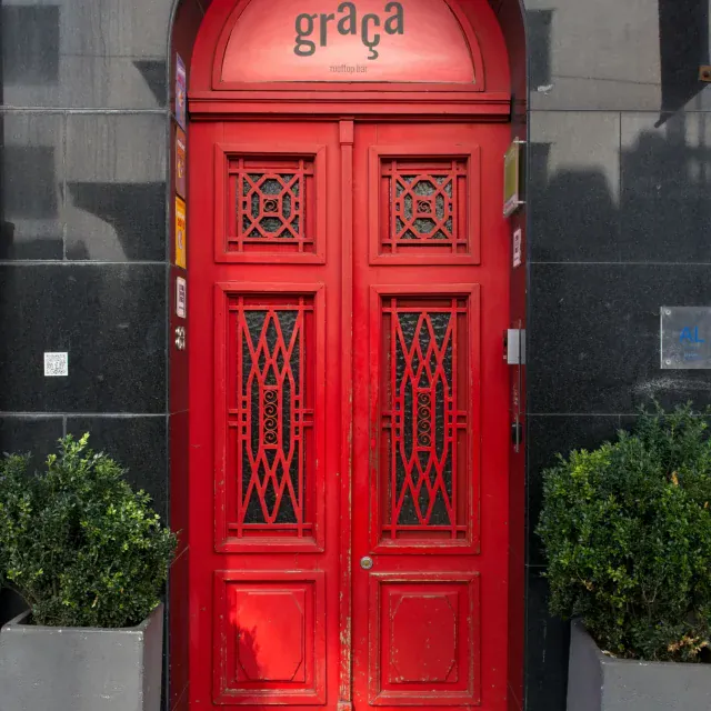 Vibrant red double door with arched top and intricate lattice grate panels, flanked by planters against a dark stone facade.