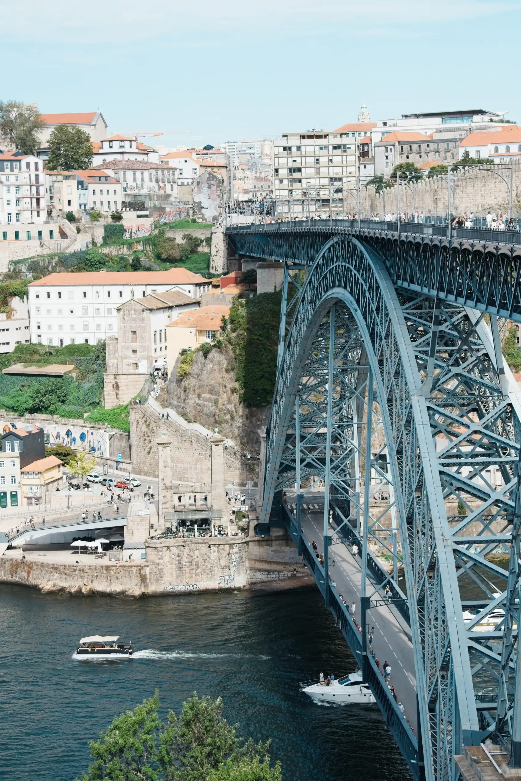 A blue arch bridge spanning a river in Porto with historic buildings on the hillside and boats below, under a clear sky.