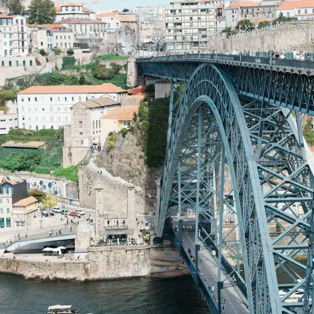 A blue arch bridge spanning a river in Porto with historic buildings on the hillside and boats below, under a clear sky.