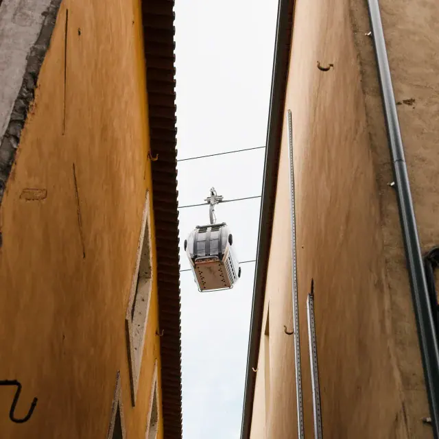 Cable car suspended between tall peach-colored buildings in a narrow alley, overhead wires visible.