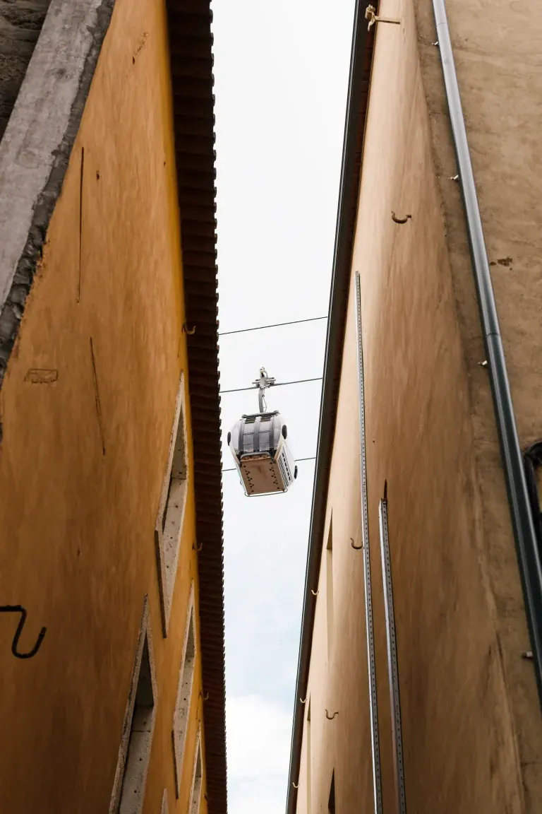 Cable car suspended between tall peach-colored buildings in a narrow alley, overhead wires visible.