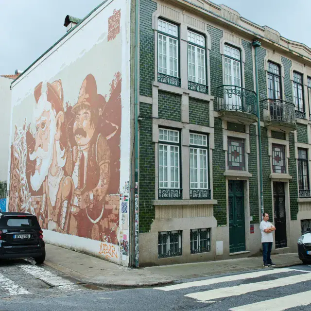 Wide street with a building facade covered by a large mural depicting vintage figures; parked cars along the curb and a person near the door.
