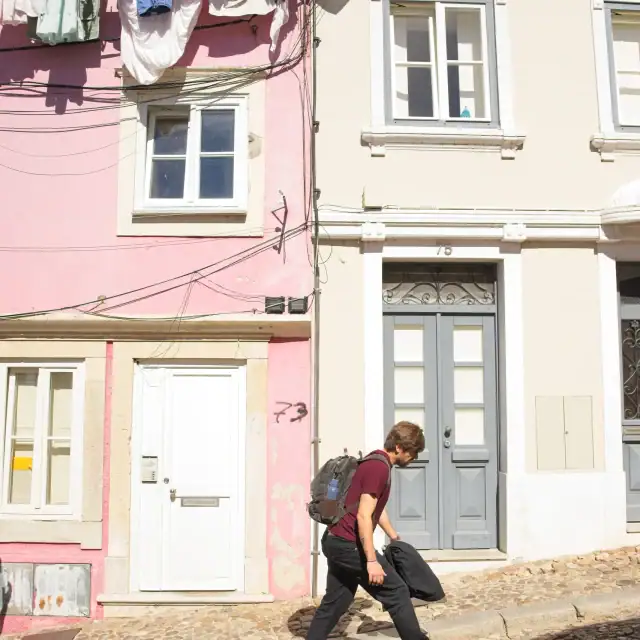 Young man walks along a cobblestone street past pastel-colored houses with laundry hanging from a line above, carrying a backpack.