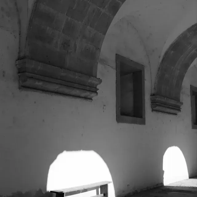 Black and white photo of a stone arcade with arches, benches, and bright openings casting light on the floor.
